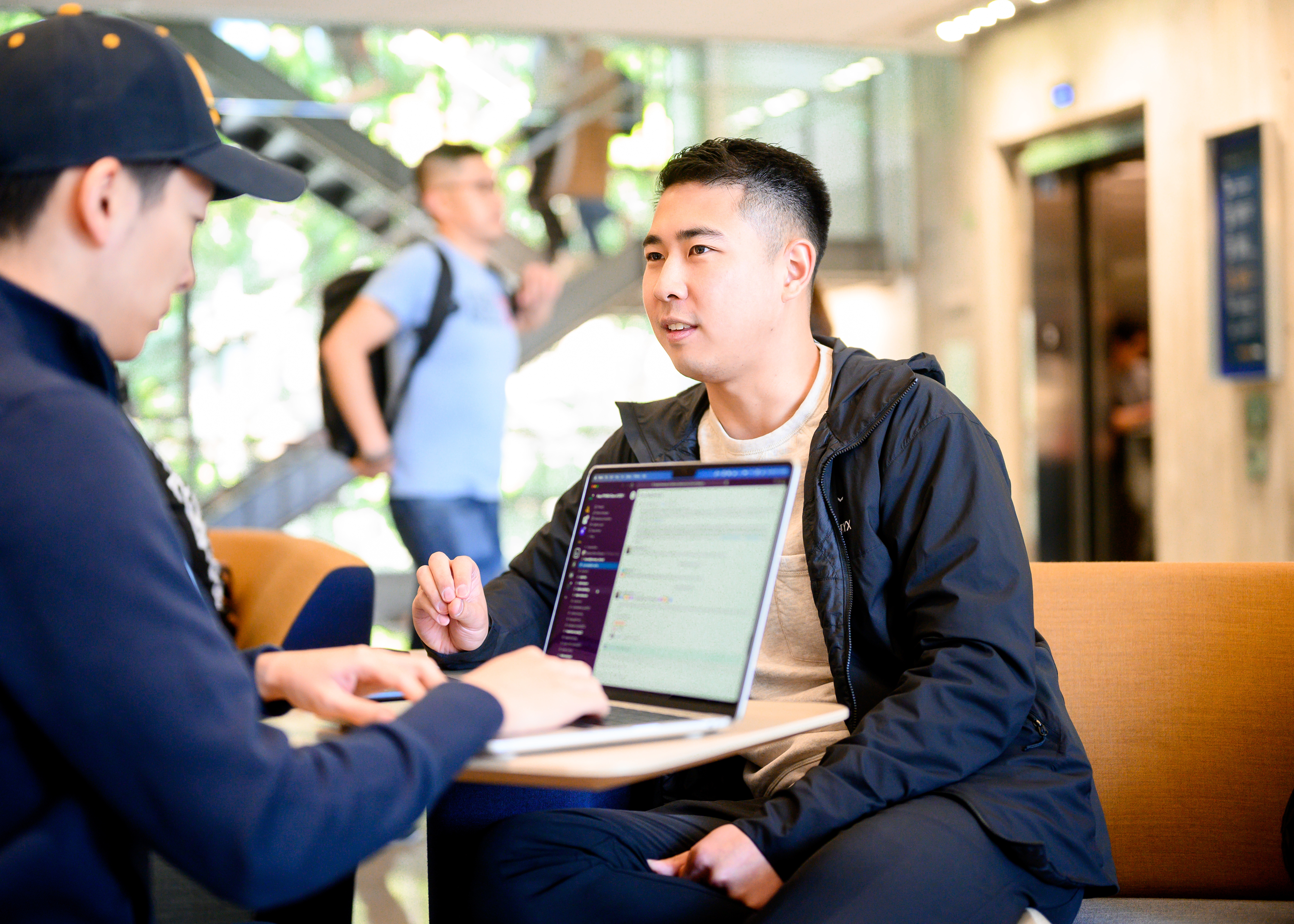 A male student speaking to another male student who is looking at a computer